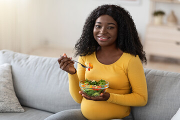 Happy pregnant black woman sitting on couch, eating fresh salad