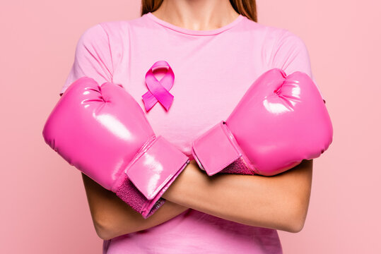 Partial view of woman in boxing gloves and ribbon of breast cancer awareness standing with crossed arms isolated on pink