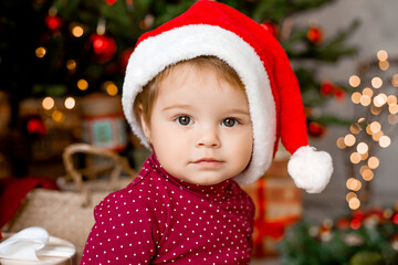 Cute baby Santa sits at home near the Christmas tree with gifts