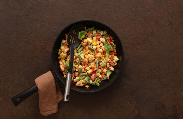 Frozen vegetables in a frying pan with a skimmer on a brown background. Copy space, top view.