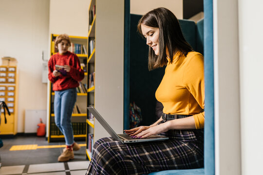 Intelligent brunette girl studying with pleasure. Female student using latop computer, among bookshelves in public library.