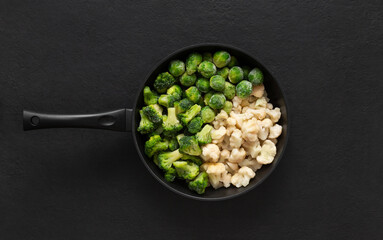 Frying pan with frozen vegetables on a black background. Cauliflower, broccoli, Brussels sprouts. Cabbage mix. Top view, flat lay.