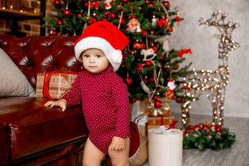 Cute baby Santa sits at home near the Christmas tree with gifts