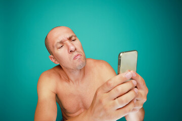 A funny bald and naked man with emotions holds mobile phone isolated on green background