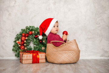 Sweet baby Santa sits in a wicker basket with a gift box and a Christmas wall on a gray background. Christmas concept, text space