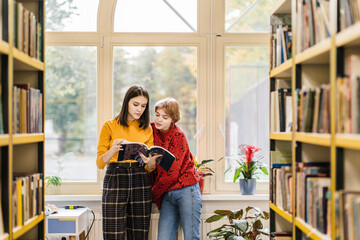 Two girls university students in library discussing plot of a novel, standing between bookshelves...