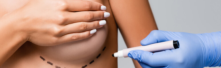 Cropped view of doctor holding marker near naked woman with marks on breast isolated on grey, banner