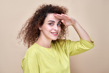 Portrait curly-haired Armenian girl dressed in green casual looking far away and smiling with hand over head. Searching concept isolated in studio background