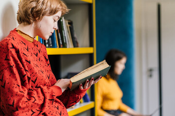 University student reading book in the library lean on on bookshelves. Young female with short hair in red sweater studying in book in library.