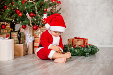 Cute baby Santa sits at home near the Christmas tree with gifts