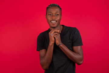 Dreamy charming Young African American handsome man standing against red background with pleasant expression, keeps hands crossed near face, excited about something pleasant.