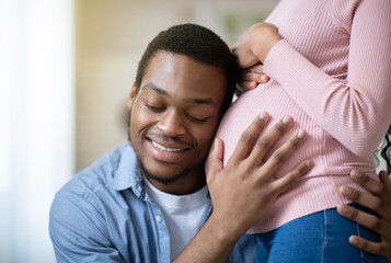 Fototapeta premium Young black man pressing his ear against his wife's pregnant belly, listening to his baby's heartbeat indoors