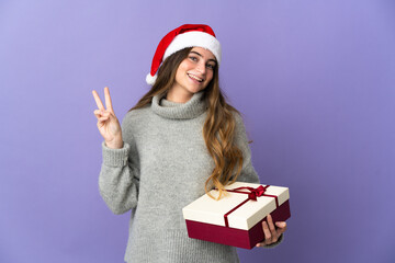 Girl with christmas hat holding a present isolated on white background smiling and showing victory sign