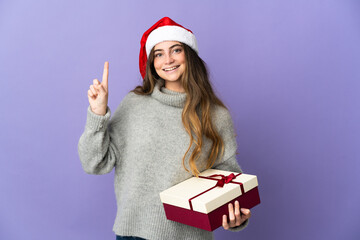 Girl with christmas hat holding a present isolated on white background showing and lifting a finger in sign of the best