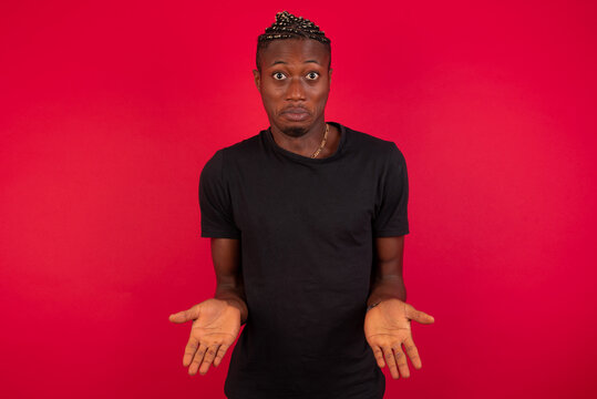 Confused Young African American Handsome Man Standing Against Red Background Chooses Between Two Ways, Points At Both Sides With Crossed Hands, Feels Doubt. Need Your Advice.