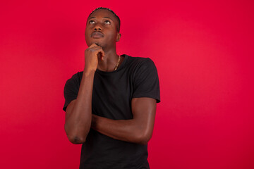 Young African American handsome man standing against red background holding hand under chin and looking upwards. Taking decisions concept. Copy-space. 