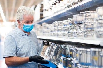 Obraz premium A white haired senior man looking at household steel pans in a store wearing a surgical mask due to the coronavirus covid-19. Consumerism concept