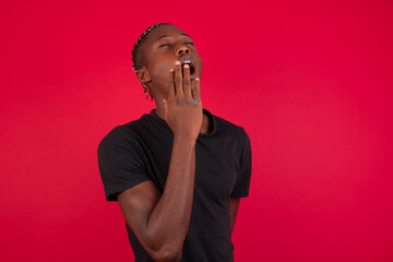 Sleepy Young African American handsome man standing against red background yawning with messy hair, feeling tired after sleepless night, yawning, covering mouth with palm.