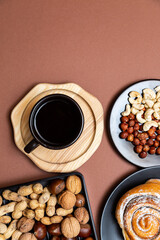 Healthy breakfast, cinnamon bun, cashews, hazelnuts, walnuts, chestnuts and peanuts on black plates with a cup of black coffee on a brown background. Place for text.