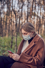Fototapeta premium A charming girl in a medical protective mask with a phone in her hands is sitting on a bench in a Park