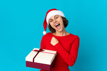 English girl with christmas hat holding a present isolated on blue background celebrating a victory