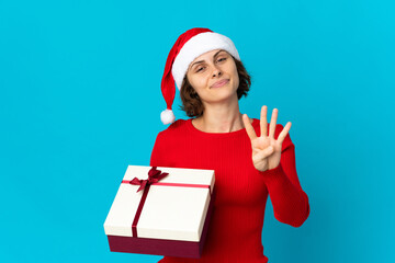 English girl with christmas hat holding a present isolated on blue background happy and counting four with fingers