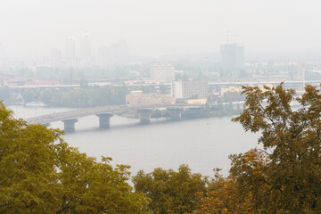 Fototapeta premium Kyiv, Ukraine-November 03, 2020:Autumn foggy morning landscape view of Dnipro River with The Harbour Bridge. Building in morning mist on the horizon. Tree Leaves Border. Natural Frame