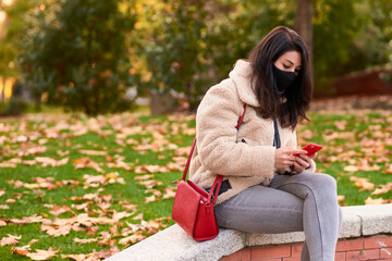 Beautiful woman with facemask sending a text message with her smartphone in a park