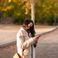 Beautiful woman with facemask sending a text message with her smartphone in a park