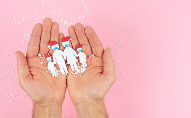 Hands holding a family cut out of paper with mask and Christmas hat on a pink background. Copy space.