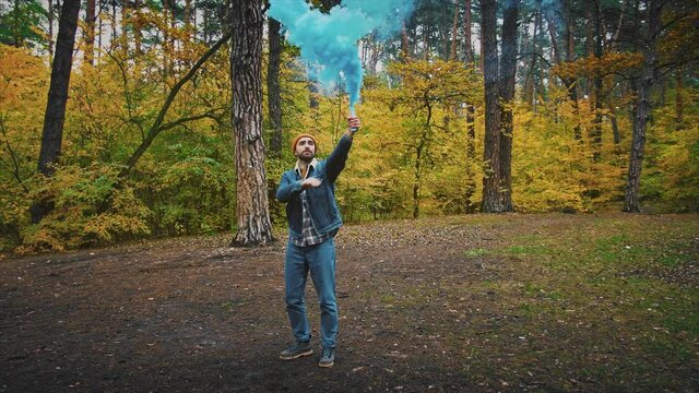 Young man in forest having fun with blue smoke grenade in autumn forest, 360 degree tracking arc shot