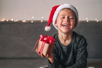 Funny smiling joyful child boy in Santa red hat holding Christmas gift in hand. Christmas concept.