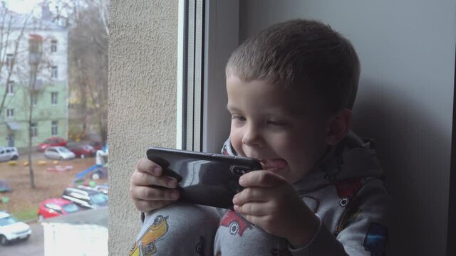 four year old boy with smartphone on windowsill. View of autumn Park and street space for text. Danger of internet
