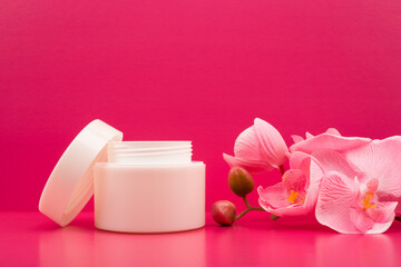 Fototapeta na wymiar Cosmetic cream in a white jar and flowers on bright pink background with space for text
