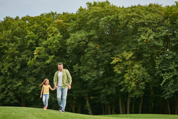 Full length shot of young father and his little daughter holding each other hands while walking in the beautiful green park on a summer day