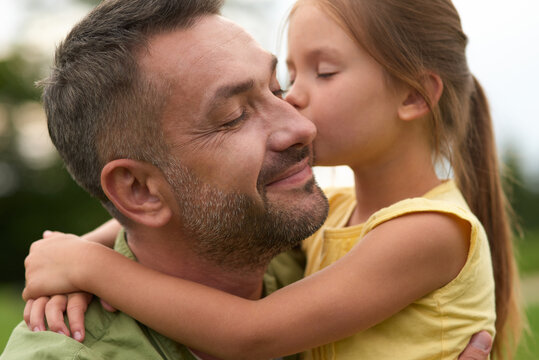 Cute little girl hugging and kissing her happy dad while spending time together outdoors on a warm day