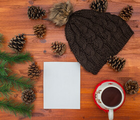 knitted hat. pine cones and green needles. cup of coffee. plain white sheet of paper. wood table. Christmas background
