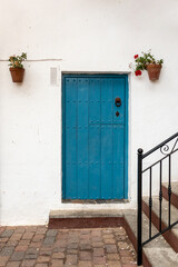 Blue wooden door on a white facade, typical house in a Spanish traditional village. Closed door with a metal handle, flower pots with geraniums hanging from the wall. Almería, Andalusia, South Spain