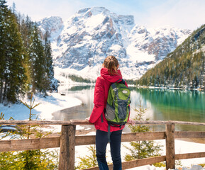 Fototapeta na wymiar Young woman with backpack near wooden fence and snowy shore of Braies lake with clear water at sunny day in winter. Travel. Landscape with slim girl, reflection in water, mountains in snow, trees