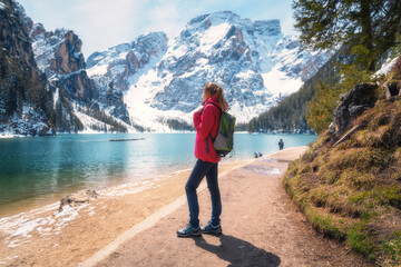 Young woman with backpack on the snowy shore of Braies lake with clear water at sunny bright day in winter. Travel. Landscape with slim girl, reflection in water, mountains, green trees, blue sky