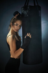 sporty brunette woman in sportswear with boxing bandages near bag on dark background with smoke.