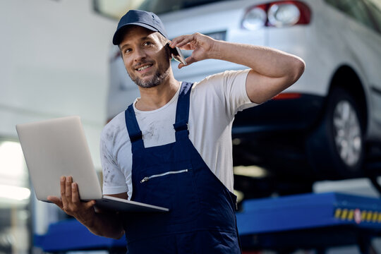 Happy Mechanic Using Computer And Talking On Cell Phone At Auto Repair Shop.