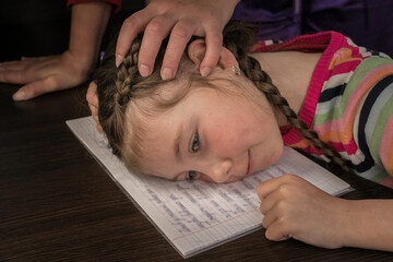 Sad little girl got tired of writing and put her head on the notebook. Mother's hand on her...