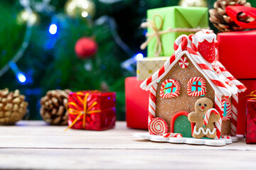 Gingerbread house on a wooden table along with christmas gifts on the background of a christmas tree