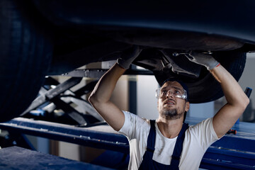 Mid adult mechanic repairing undercarriage of a car in auto repair shop. - obrazy, fototapety, plakaty