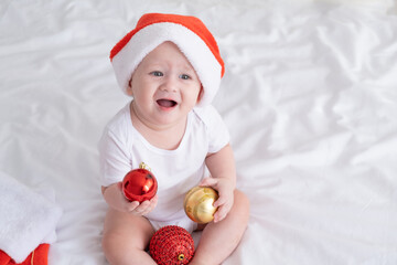 cute baby boy in Santa hat on bed with white bedding at home with Christmas decorations.