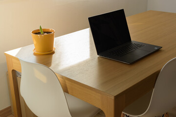 Working scene with a wooden table, design white chairs, black laptop and a small cactus in a yellow pot. Telecommuting atmosphere.