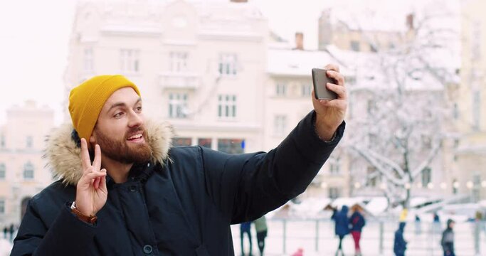 Young caucasian male tourist in yellow hat taking selfie on smartphone on winter architecture background of city.