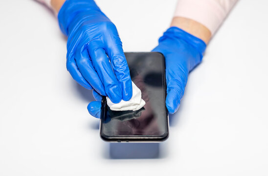 Female hand in blue protective gloves sanitizing and cleaning smartphone or mobile phone with alcohol sanitizer and wipe on white table. Top view