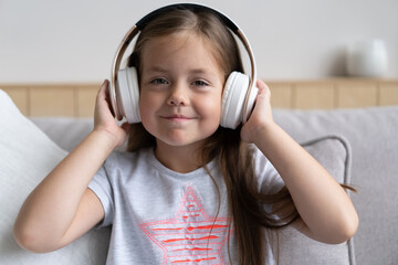 Adorable small schoolgirl sitting on sofa, wearing modern wireless headphones, listening to favorite music, Cute little child resting on couch